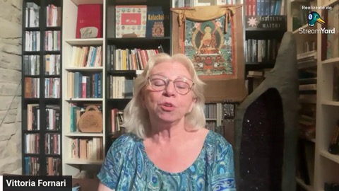 Vittoria in abito estivo a fiori verde-azzurro parla in diretta nella sua biblioteca, sotto il thangka del Buddha tra gli scaffali pieni di libri